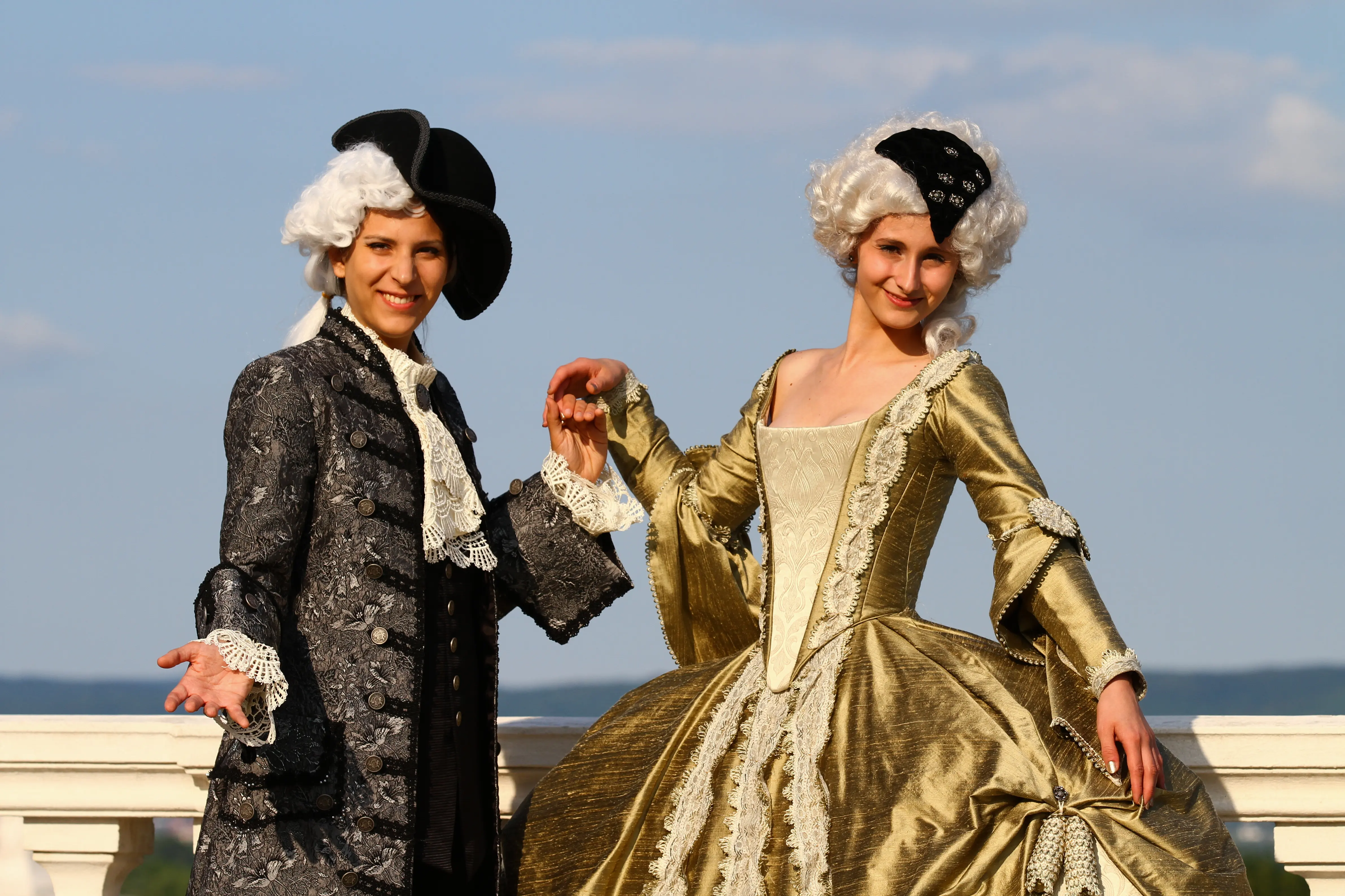 The gold gown beside a companion in a silver-grey justaucorps and tricorn on the Schloss Hof terrace under a blue sky